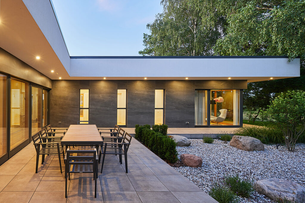 Outdoor dining terrace of Villa AJ with modern furniture, recessed lighting, dark façade cladding and landscaped courtyard garden, designed by NG Architects